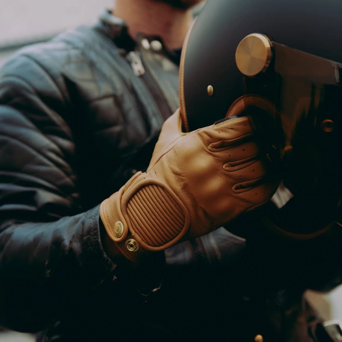 Rider wearing a tan leather glove while holding a black classic motorcycle helmet